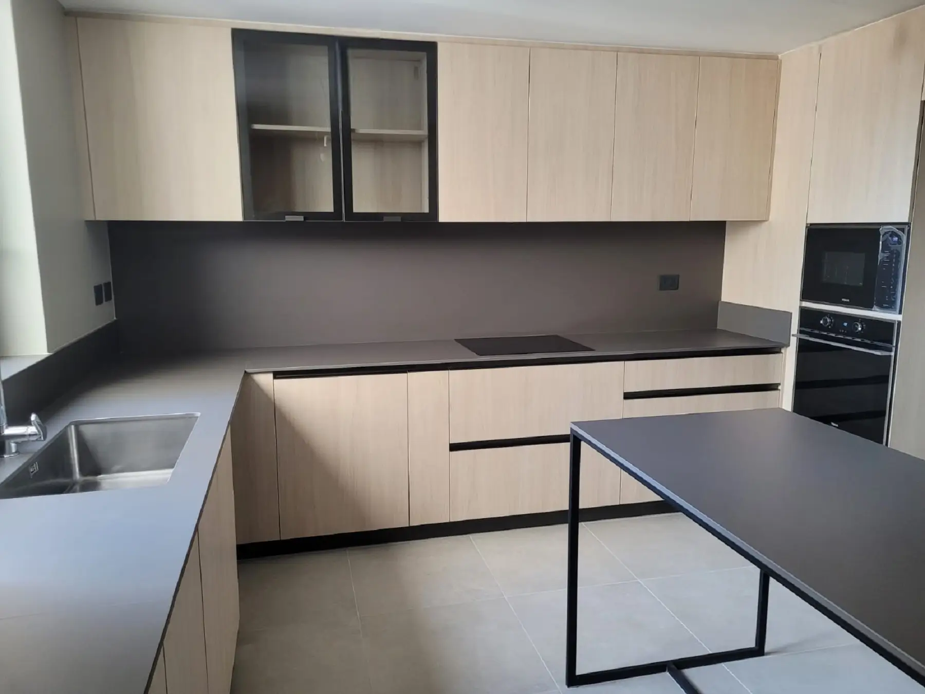 Cocina moderna fabricada en melamina en Chile con gabinetes de madera clara, cubierta de cuarzo gris oscuro y muebles superiores con puertas de vidrio