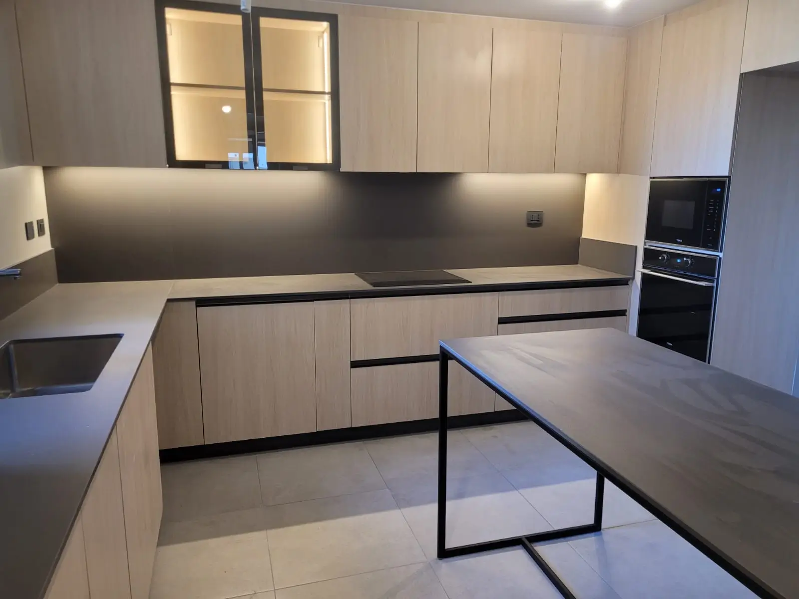 Cocina moderna fabricada en melamina en Chile con gabinetes de madera clara, cubierta de cuarzo oscuro y muebles superiores con puertas de vidrio iluminadas